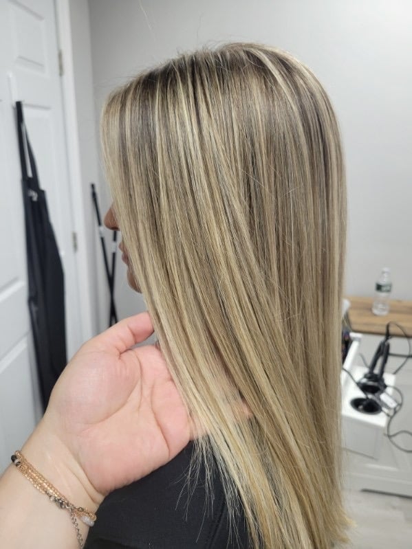 Person displaying long, straight, layered blonde hair with highlights, shown from behind in an indoor salon setting