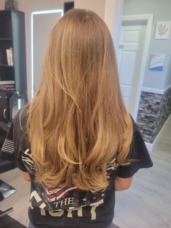 Back view of a person with long, wavy golden-brown hair wearing a black graphic t-shirt in an indoor salon or studio setting