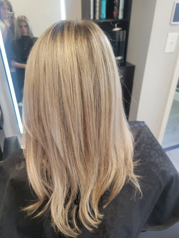 Rear view of a woman with long, straight, blonde hair in a salon setting with a mirror and professional styling cape